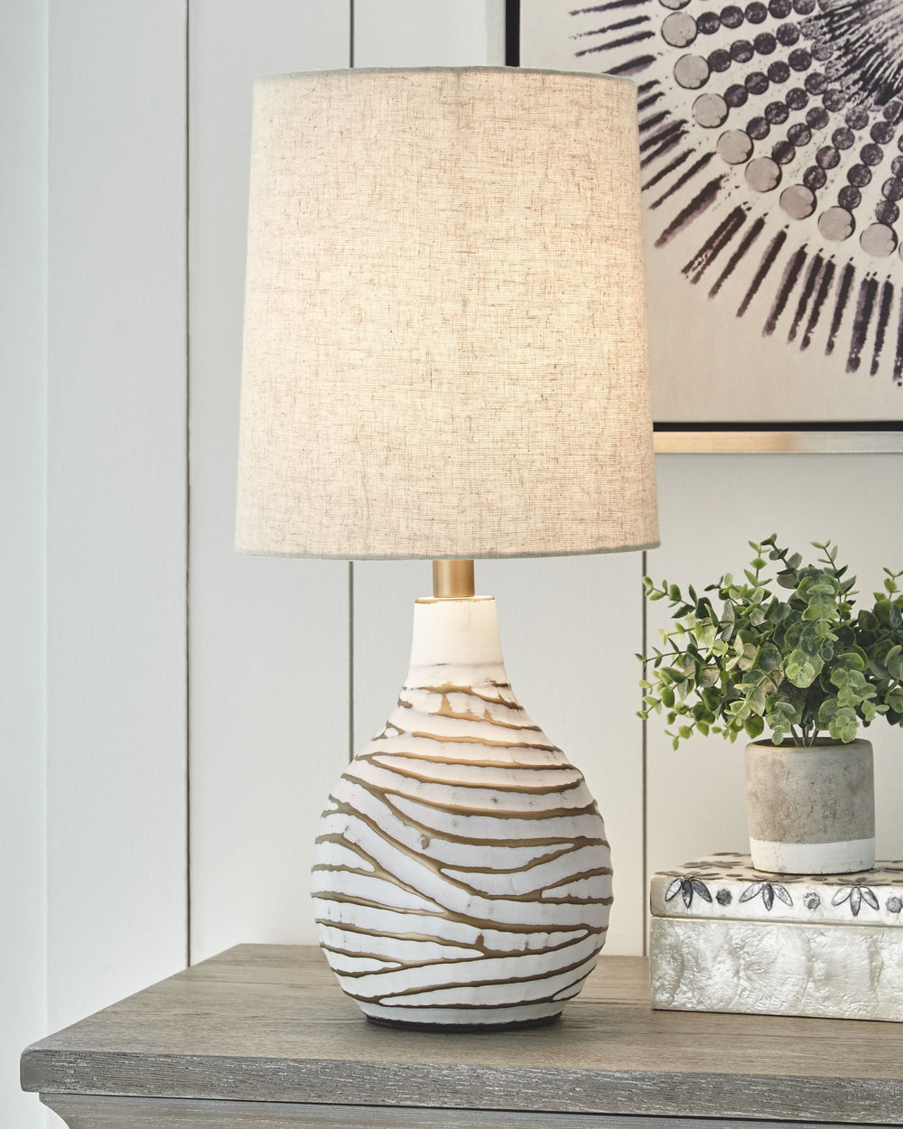 Shine a contemporary spotlight on your good taste with this table lamp. The antiqued white and goldtone finish of the metal lamp adds texture and interest to any space. With a modified drum shade, this light will illuminate your decor with style.<br><br><